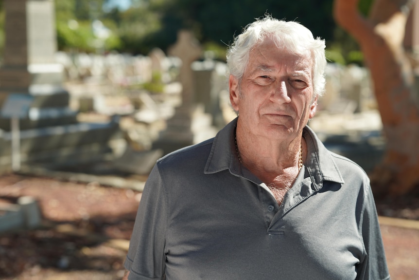 A man with light grey hair stands for portrait, cemetery in background