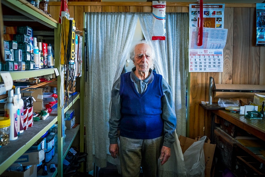 An elderly man in a blue vest and grey top stands in front of a curtain staring at the camera 