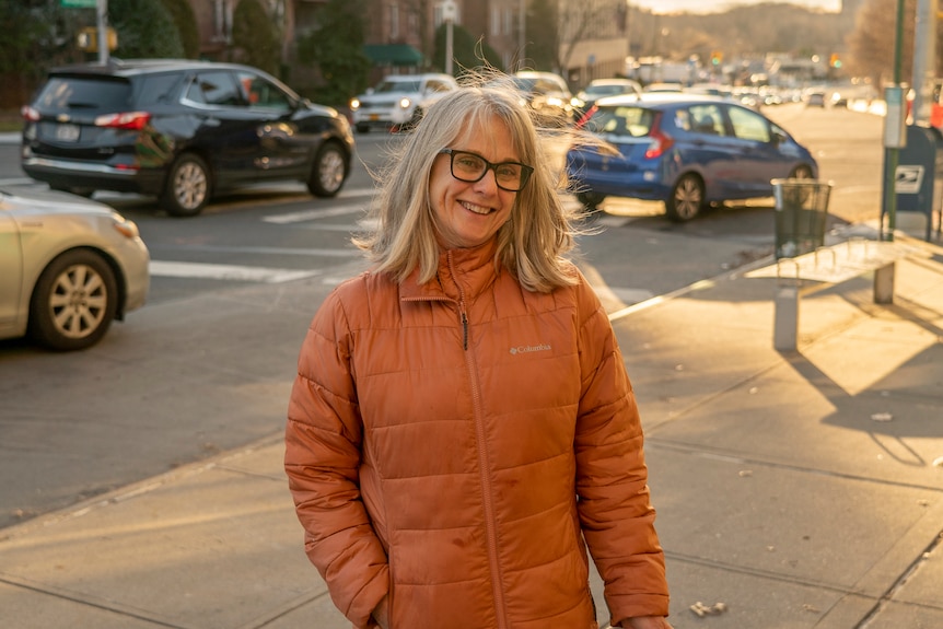A woman with black glasses in an orange puffer jacket smiles at the camera