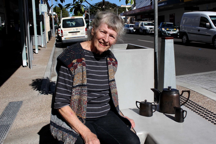 A lady is seated beside a bronze sculpture of a tea set in Adelaide Street Maryborough