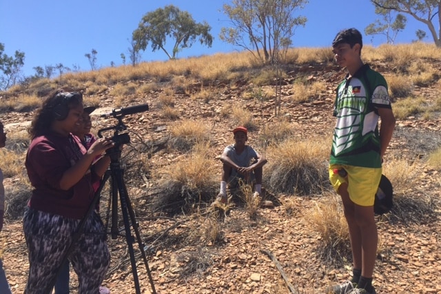 An indigenous teenage girl behind a camera and tripod films an indigenous teenage boy on a hill in Mount Isa.