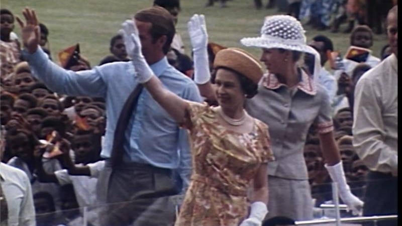 Queen Elizabeth waves while standing on motorcade, Papua New Guineans behind her.