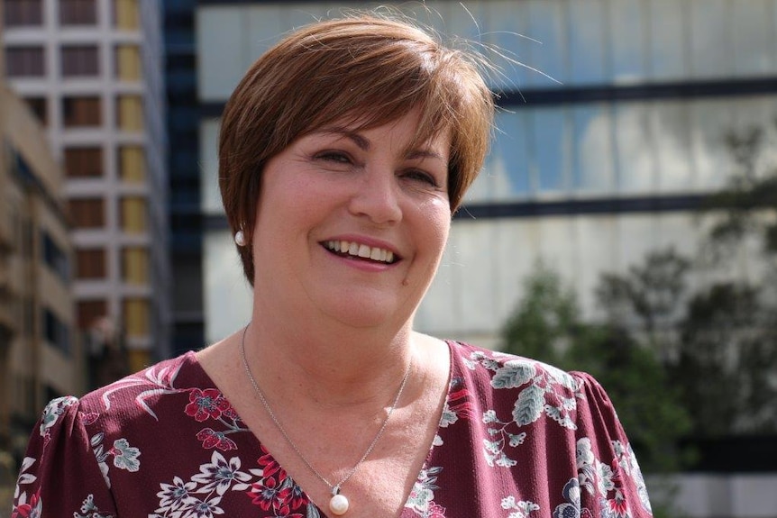 A close up of Marion wearing a dark red floral shirt, smiling at the camera with buildings in the background.