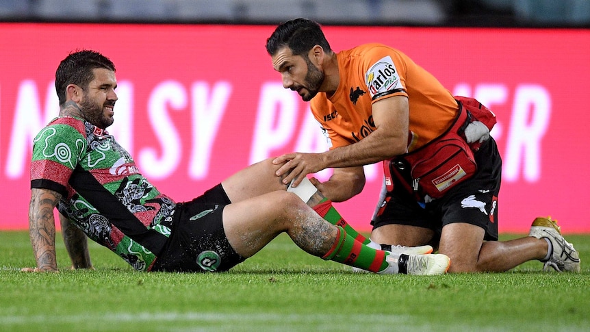 Adam Reynolds lies on the ground as a Rabbitohs trainer attends to his left knee.