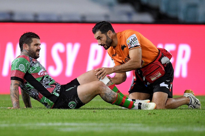 Adam Reynolds lies on the ground as a Rabbitohs trainer attends to his left knee.