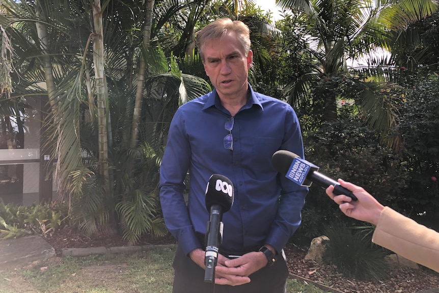 A man with grey hair and a blue shirt stands outdoors in front of two microphones.