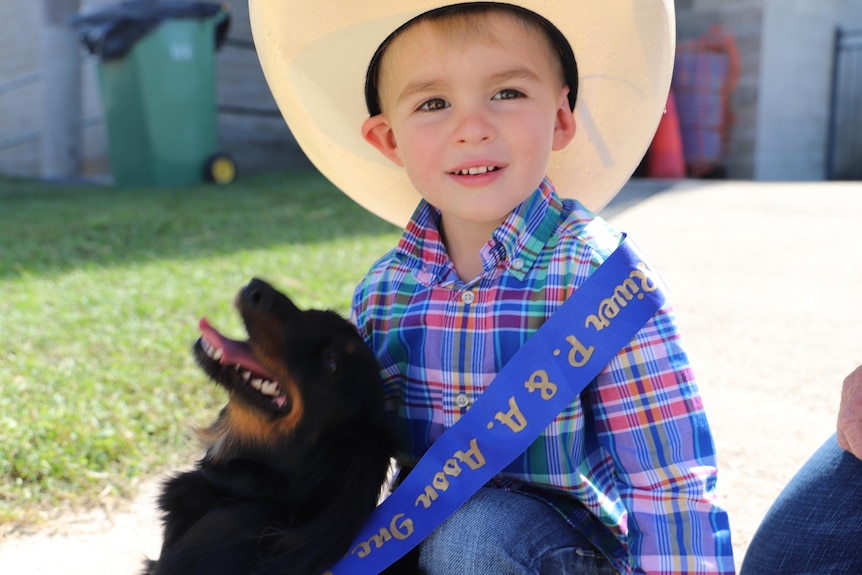 A boy wearing a cowboy hat and a blue sash cuddling into his dog.