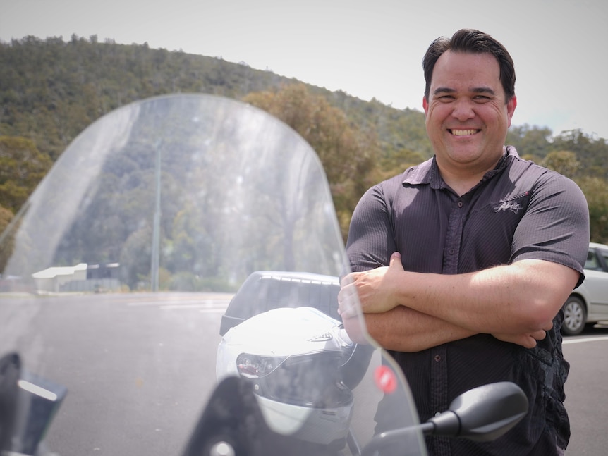 A man in a polo shirt smiling at the camera with his arms crossed, with a motorcycle windshield and rear view mirror in the foreground.