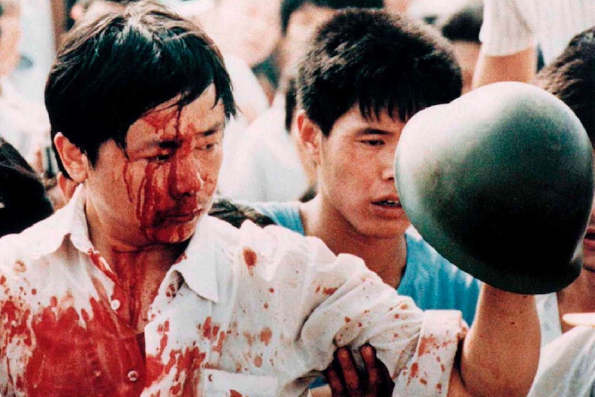 A blood-covered man in a white shirt holds a Chinese soldier's helmet in a crowd of civilians.