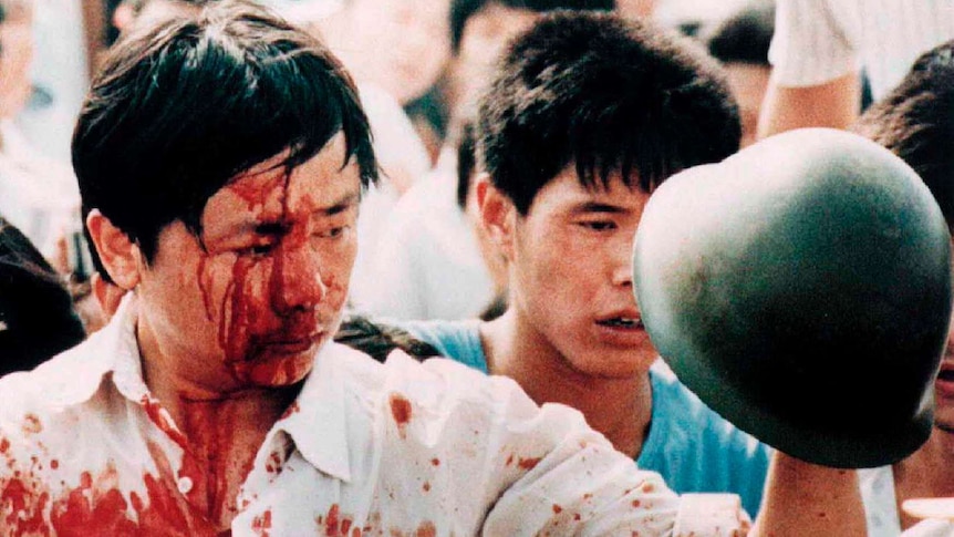 A blood-covered man in a white shirt holds a Chinese soldier's helmet in a crowd of civilians.