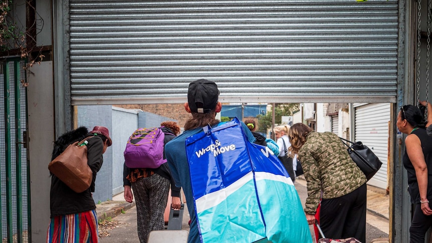 Un grupo de personas, cargando bolsas, sale de un refugio para personas sin hogar a través de una puerta enrollable.