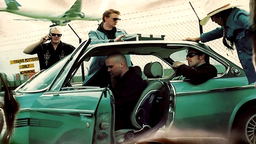 queens of the stone age stand around a vintage green car near an airport runway. josh homme has a briefcase on the room