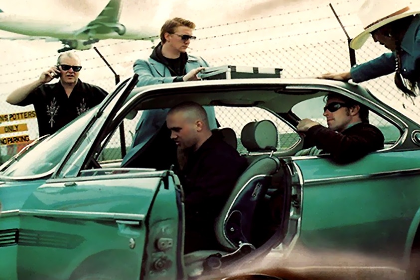 queens of the stone age stand around a vintage green car near an airport runway. josh homme has a briefcase on the room
