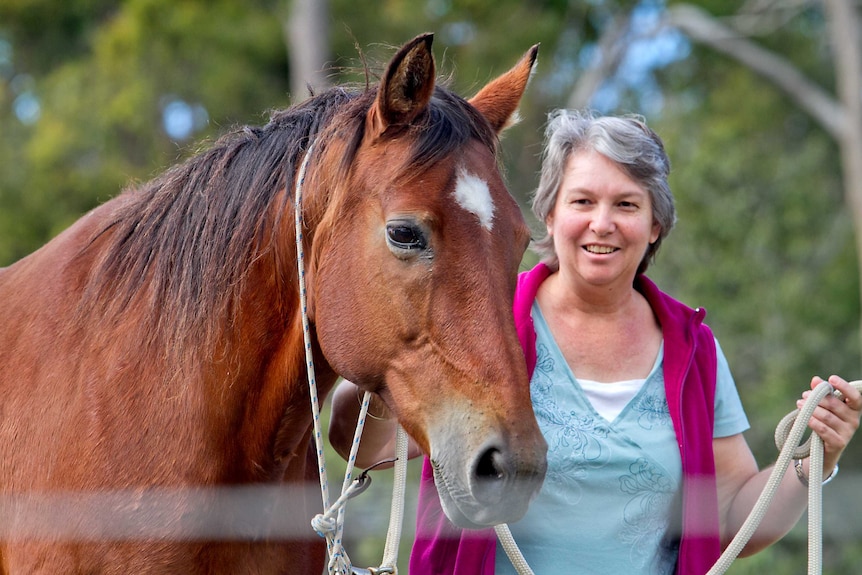 Sandra Goldner wears a magenta vest over a light blue long sleeved shirt as she stands with a brown horse