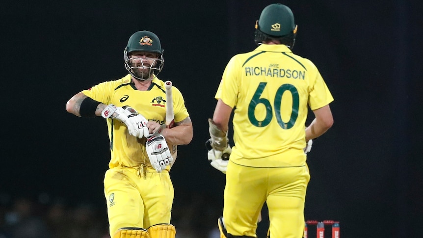 A smiling Australian T20 batsman walks down the pitch towards his teammate while taking his gloves off after the game. 