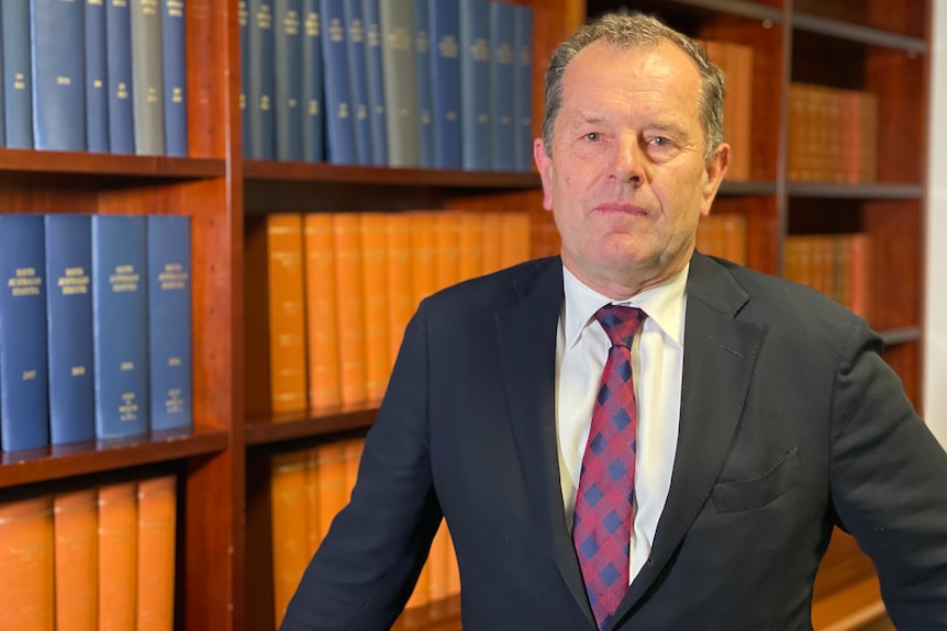 A man in a black suit, white shirt and red and blue tie stands in front of a bookcase holding leather-bound books