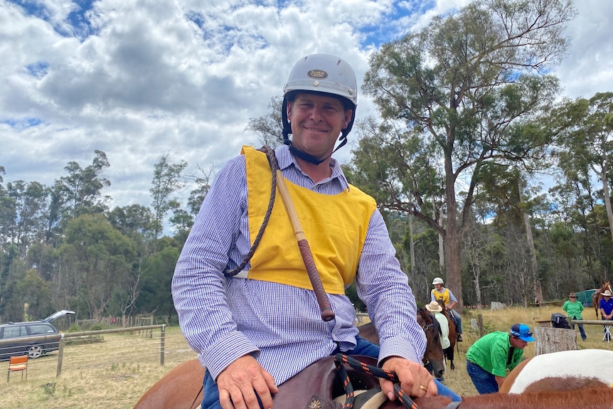 A man in a white helmet, blue shirt and yellow bib sits on a chestnut horse, he has a stock whip over his shoulder. 