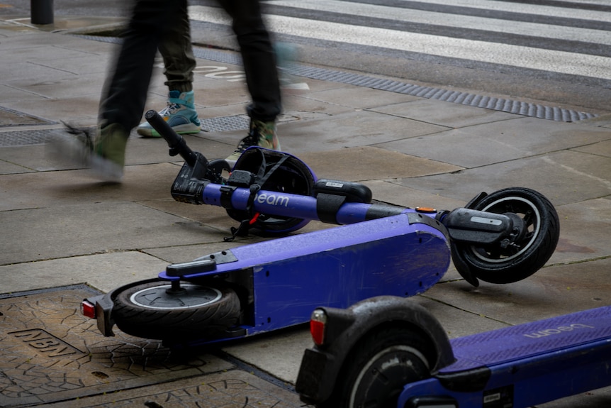 Dos e-scooters morados de costado en un sendero, con dos pares de piernas caminando