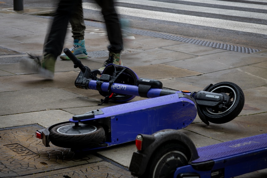 Two purple e-scooters on their sides on a footpath, with two pairs of legs walking past