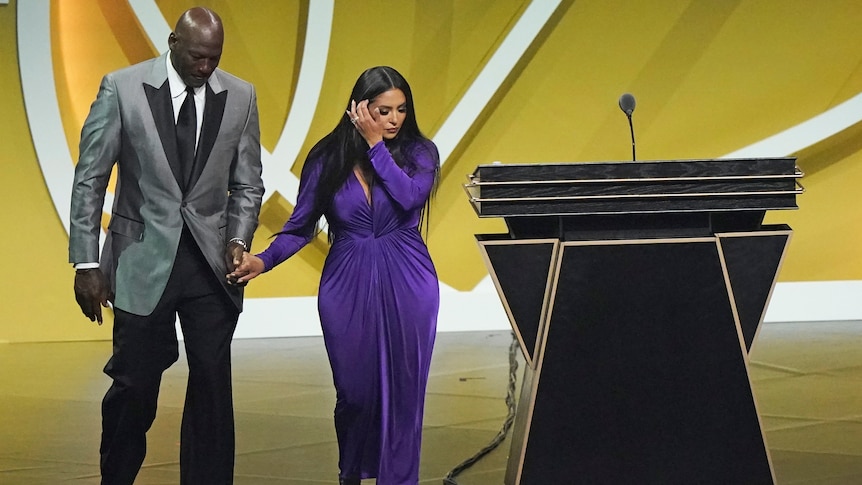 Michael Jordan and Vanessa Bryant are emotional as they walk off a stage holding hands. He is in a suit and she a purple dress