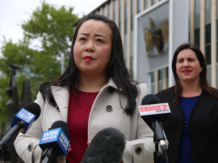 Elizabeth Lee stands in front of microphones while Giulia Jones stands in the background.