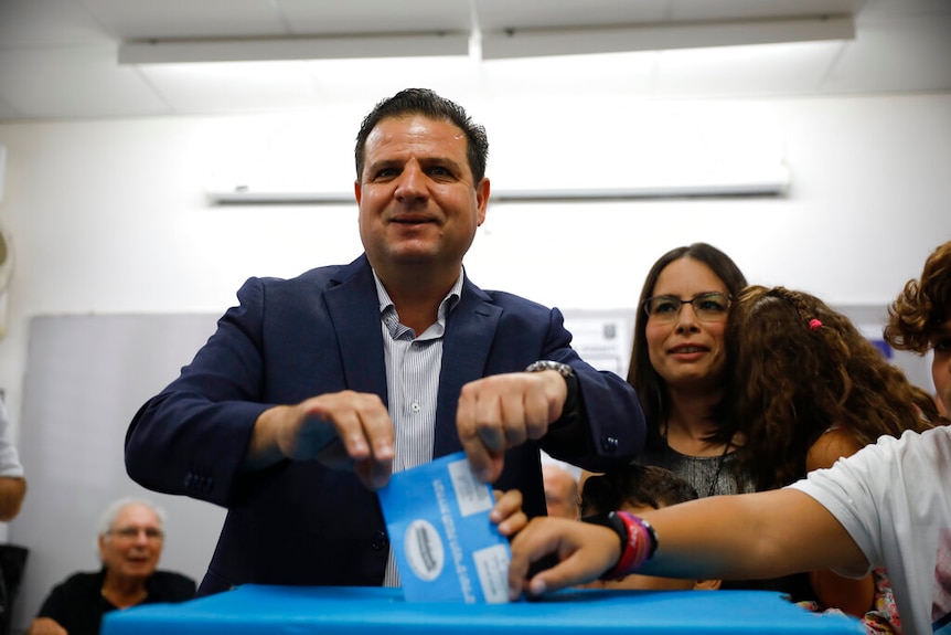 Israeli Arab politician Ayman Odeh holds his blue voting envelope over a blue ballot box as a woman and baby look on.