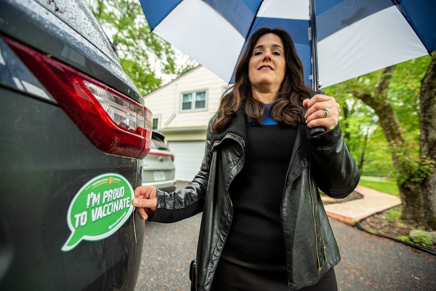 A woman holding an umbrella puts a sticker reading "I'm proud to vaccinate" on the back of her car