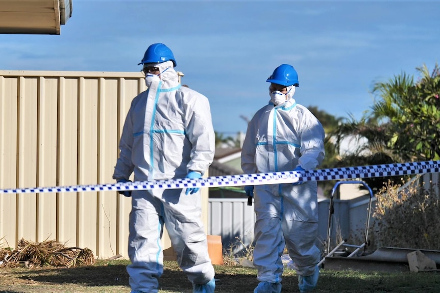 Two people in white coveralls, blue helmets and face mass walk in yard 