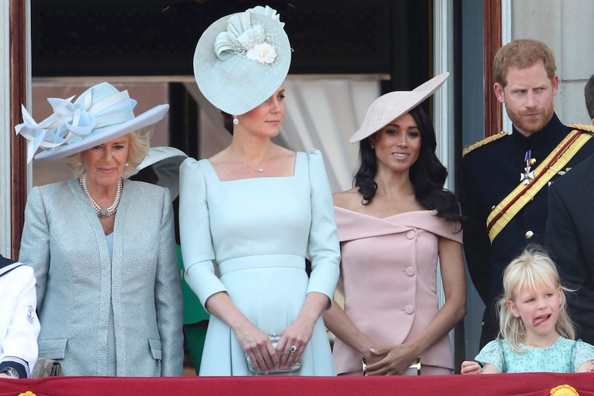 Three well dressed women, one man with a sash and a girl with her tongue out stand behind bunting