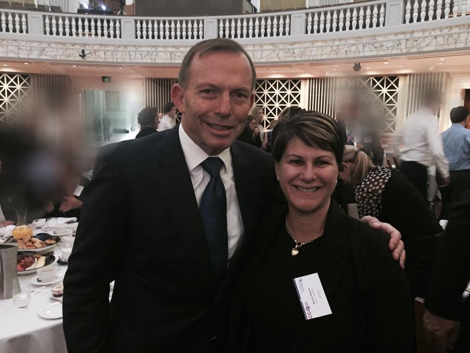 A woman stands with tony abbott in a large room in front of a table of food