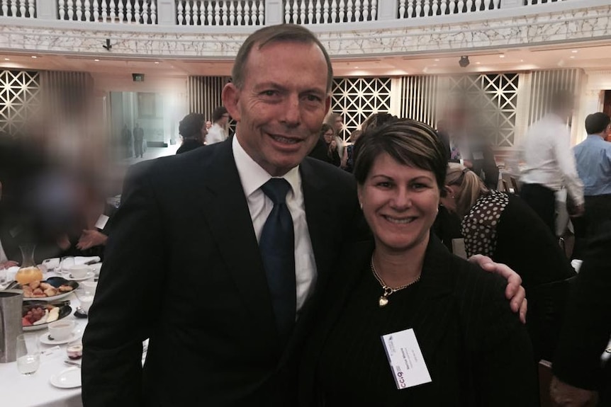 A woman stands with tony abbott in a large room in front of a table of food