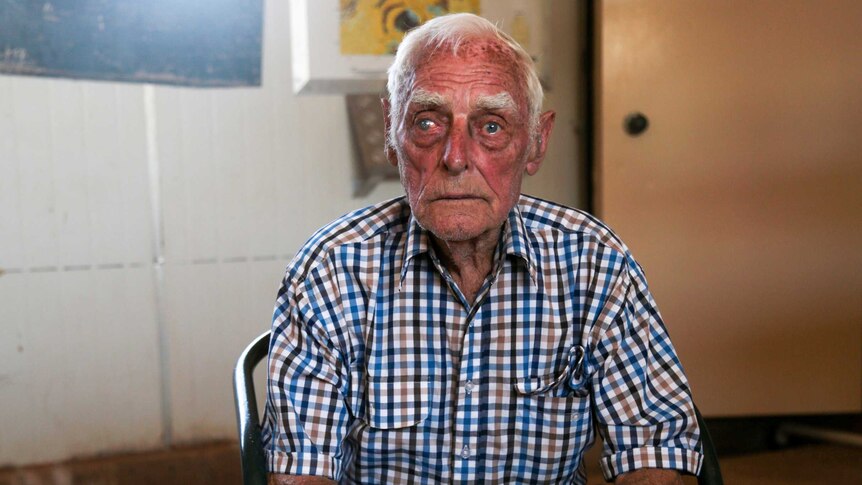 An old man wearing a checked shirt sits on a chair with his hands clasped in front of him