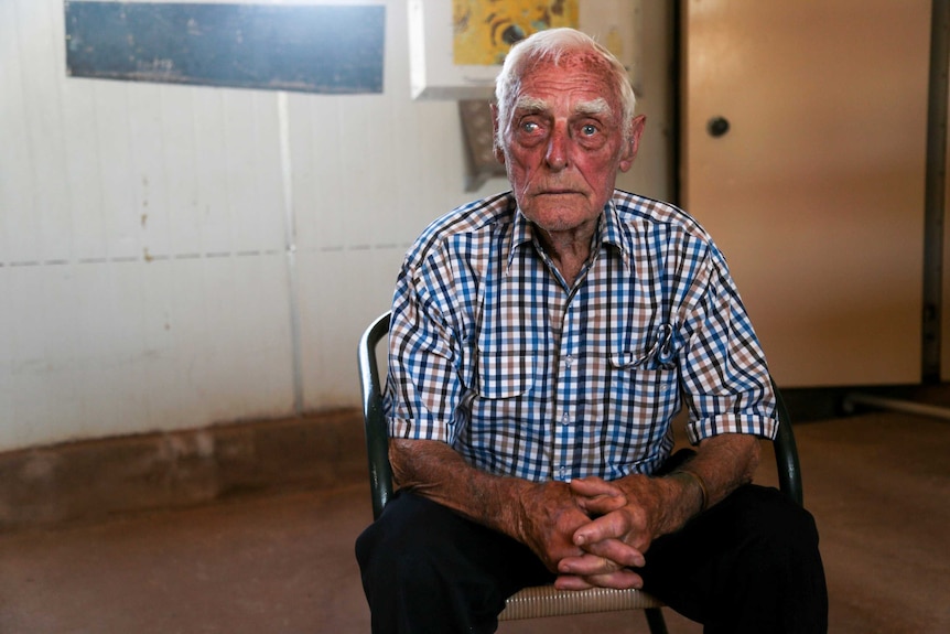 An old man wearing a checked shirt sits on a chair with his hands clasped in front of him