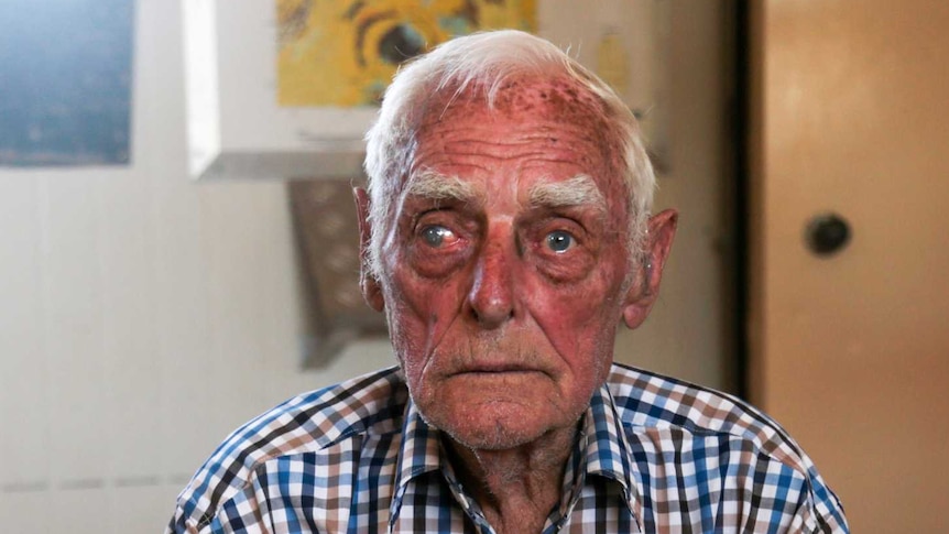 An old man wearing a checked shirt sits on a chair with his hands clasped in front of him