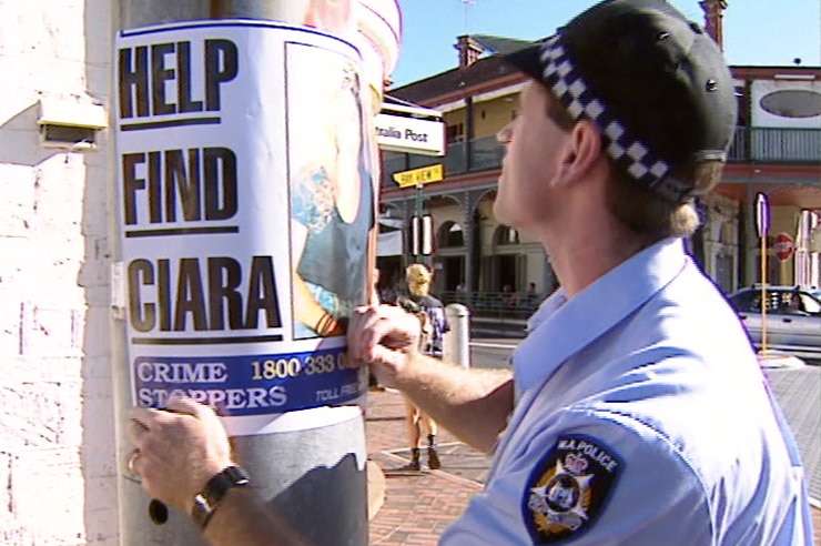 A policeman wearing a cap tapes a "Help Find Ciara" poster to a light pole