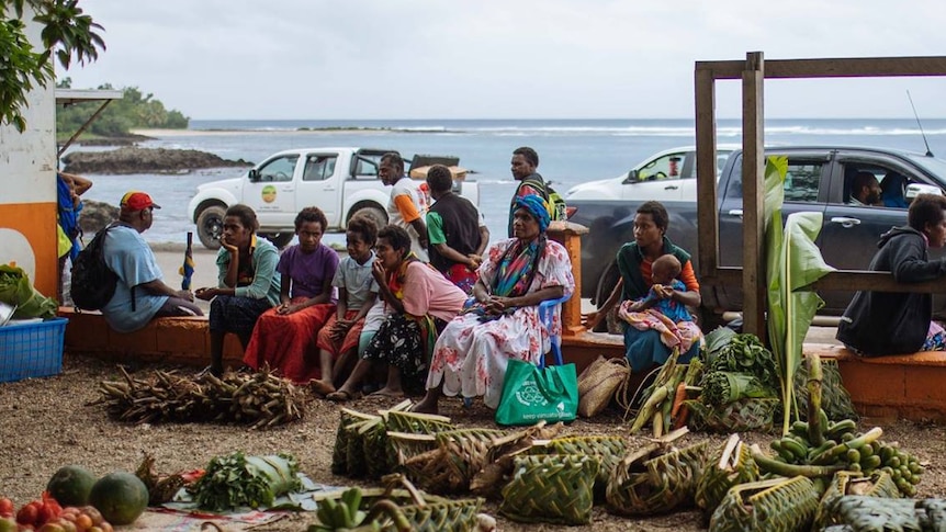 Women sit along a fence at a fruit and vegetable market on an overcast day with the ocean in the background 