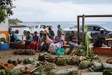 Women sit along a fence at a fruit and vegetable market on an overcast day with the ocean in the background 