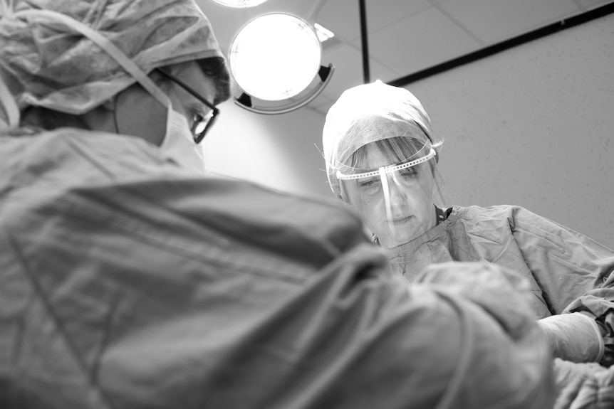 A woman wearing PPE looks down under a bright light in a surgical setting.