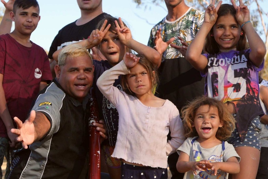 An Aboriginal man kneels holding a didgeridoo, surrounded by several Aboriginal children smiling at the camera.