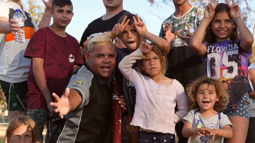 An Aboriginal man kneels holding a didgeridoo, surrounded by several Aboriginal children smiling at the camera.