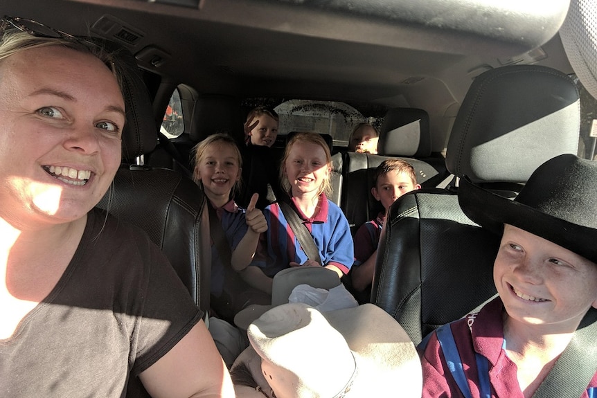 A woman takes a selfie of her and six kids in school uniform in a car with their seatbelts on