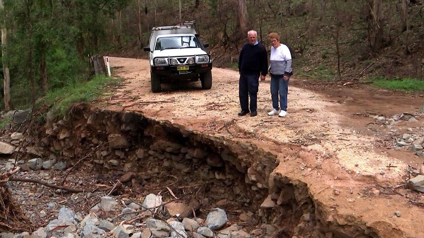 An elderly couple inspect damage to a dirt road which has left a large crater.