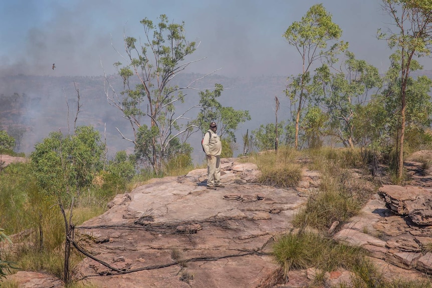 a man on a cliff with a fire in the background