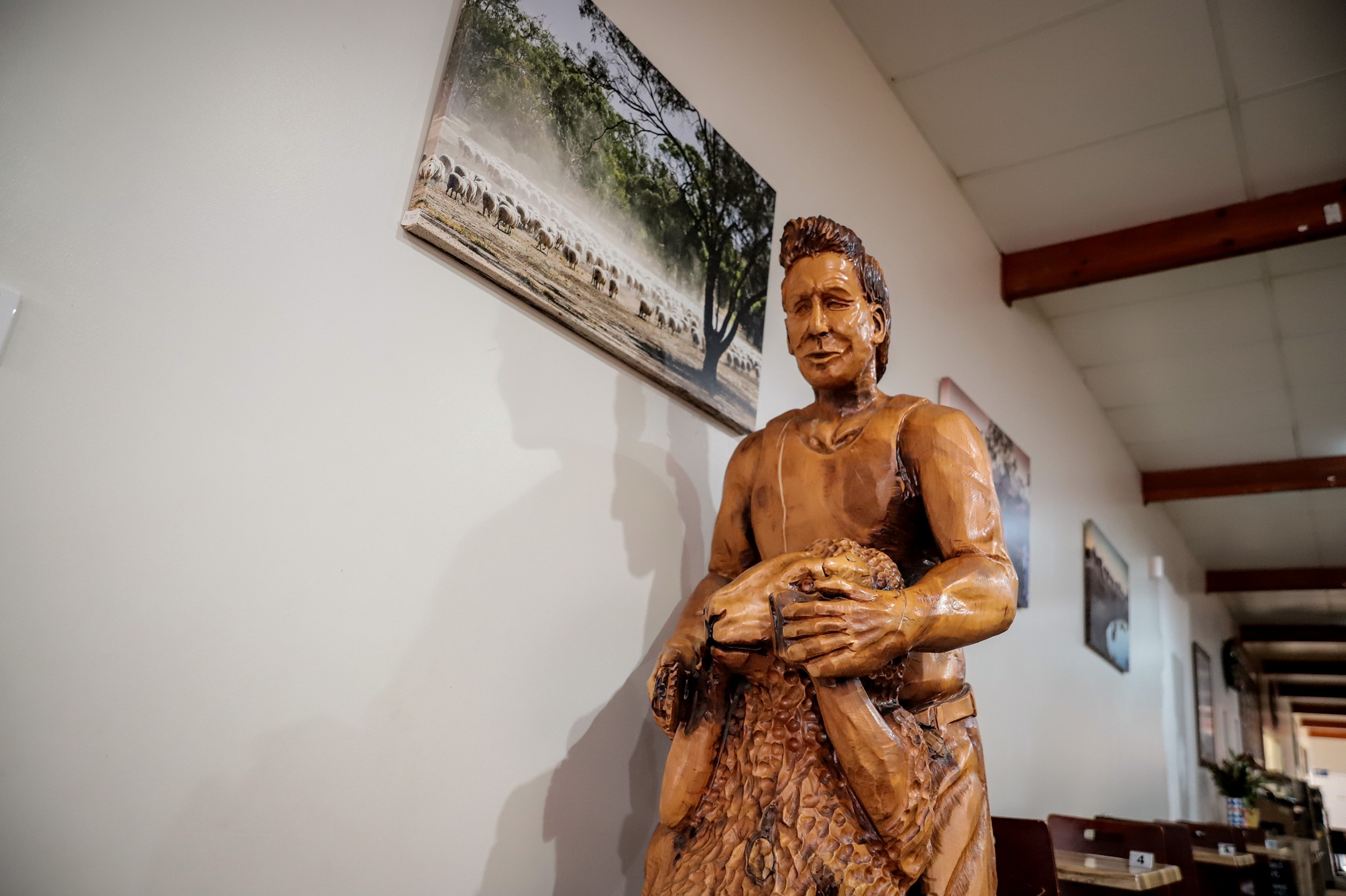 A carved wooden statue of a man wearing a singlet, holding a sheep is seen in a room with photos of farming scenes on the wall