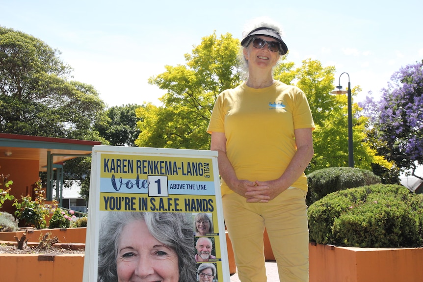 A woman dressed in yellow stands next to an election corflute.