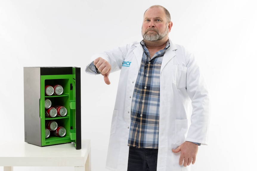 A man in a lab coat standing in a white room next to a small bar fridge, making a thumbs down gesture