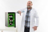 A man in a lab coat standing in a white room next to a small bar fridge, making a thumbs down gesture