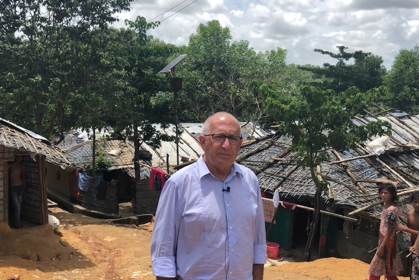 Chris Sidoti in a purple shirt stands inside the refugee camp, he is surrounded by huts constructed out of tarpaulins and bamboo
