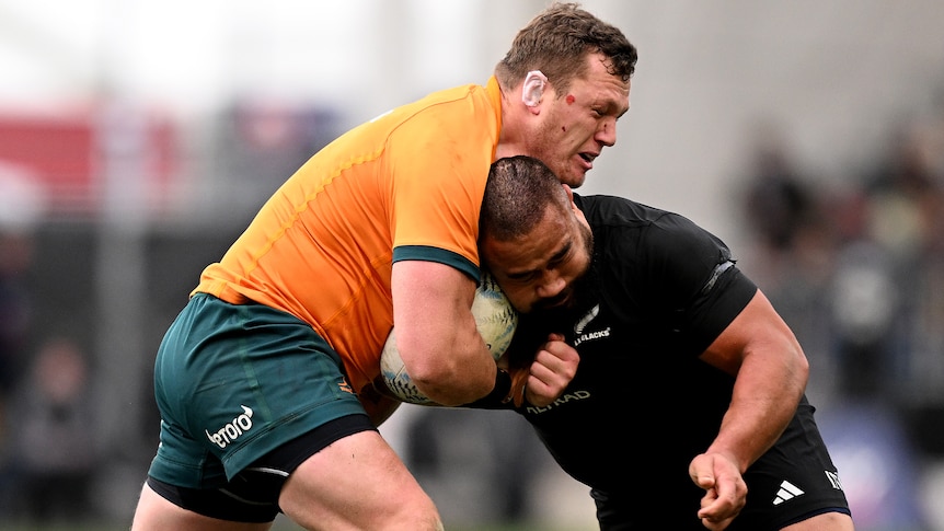 A Wallabies player holds the ball as he collides with an All Blacks opponent.