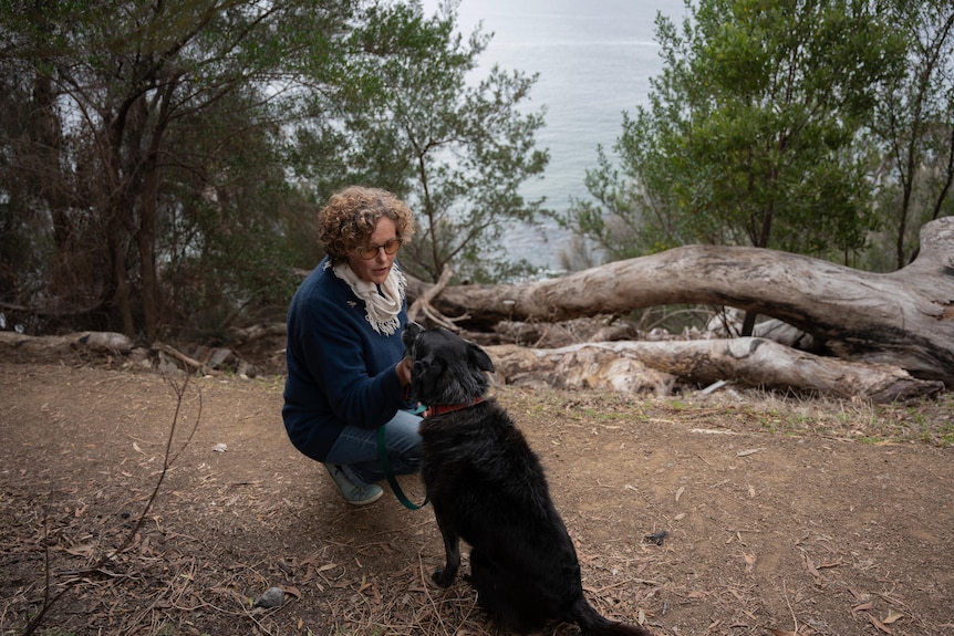 A woman kneels down on a bush track and looks into the eyes of her black dog.